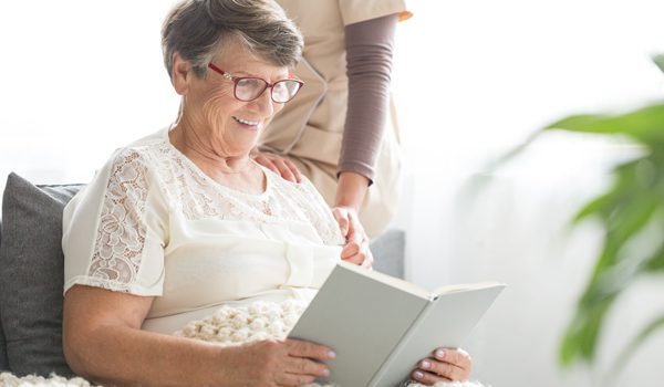 smiling-senior-patient-sitting-on-sofa-with-blanket-on-legs-and-reading-book-at-nursing-home