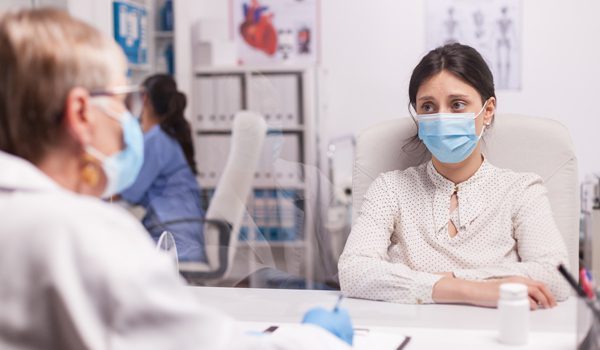 stressed-young-patient-with-face-mask-against-covid-19-during-consultation-with-senior-doctor-in-hospital-office-terrified-woman